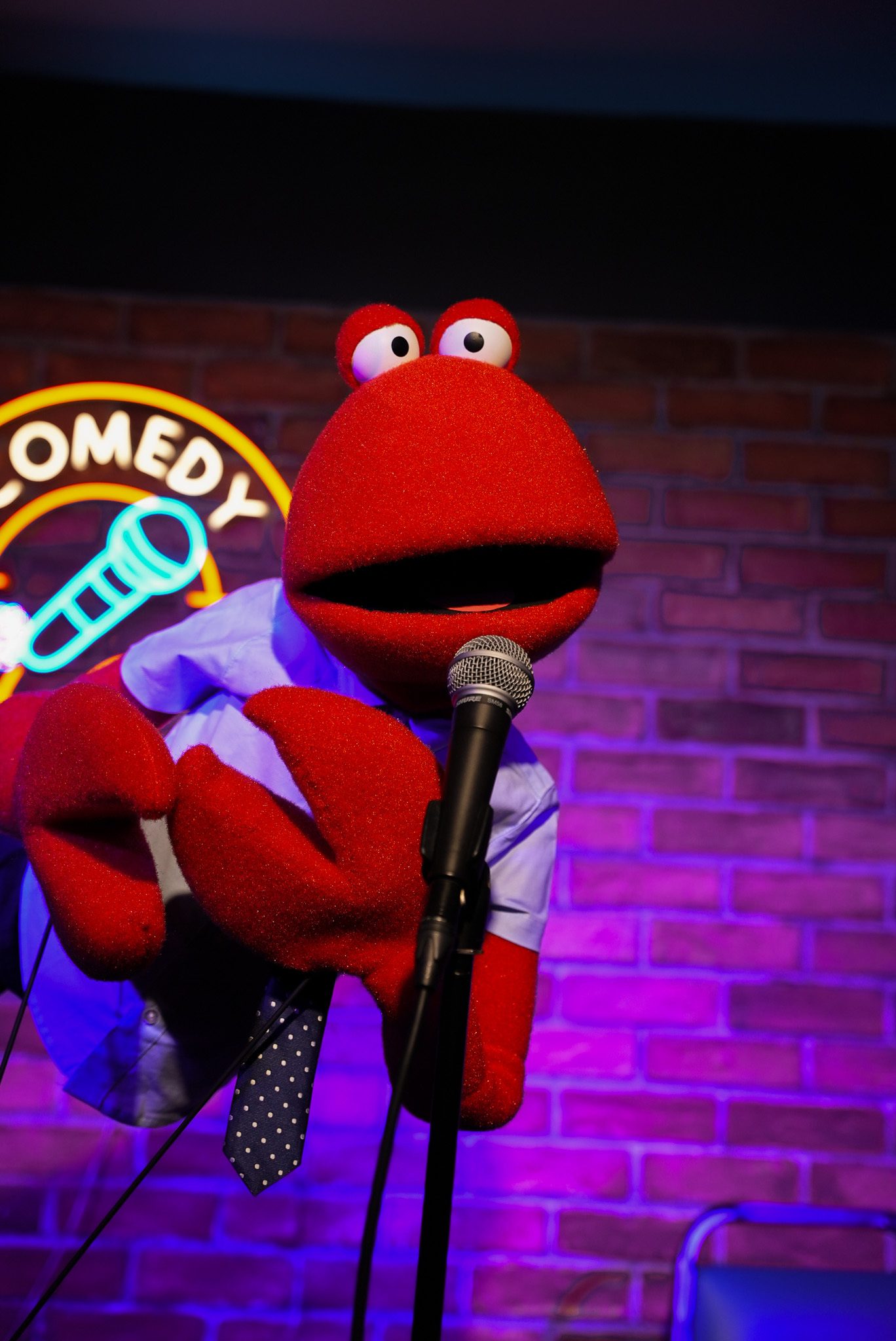 A bright red puppet with a polka-dot tie leans into a microphone on a comedy club stage with neon lighting